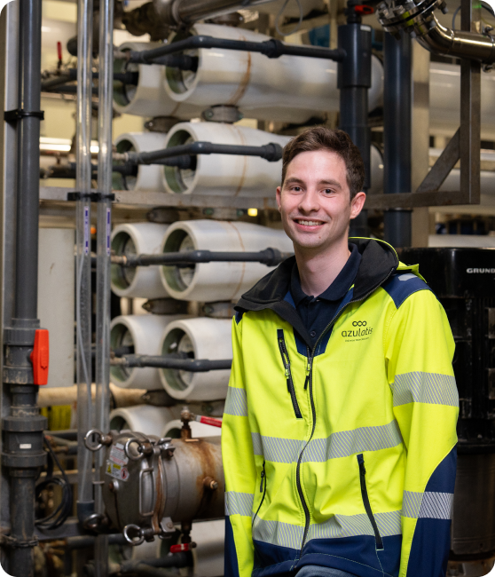 Een jonge man met kort bruin haar glimlacht naar de camera terwijl hij in een industriële omgeving staat. Hij draagt een gele veiligheidsjas met reflecterende strepen en het logo van "Azulatis". Op de achtergrond zijn metalen buizen, kleppen en filtratiesystemen zichtbaar.